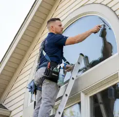 Professional window cleaner on a ladder using a squeegee to clean an exterior arched window, Long Beach, CA.